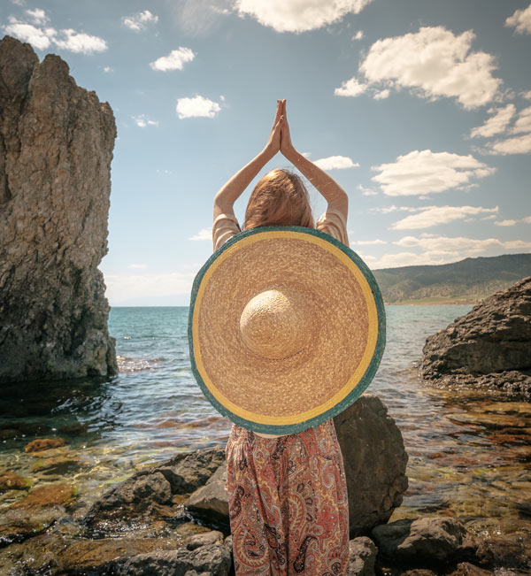 woman in a large straw hat doing yoga by the sea surrounded by rocks relaxation summer wellness mindfulness